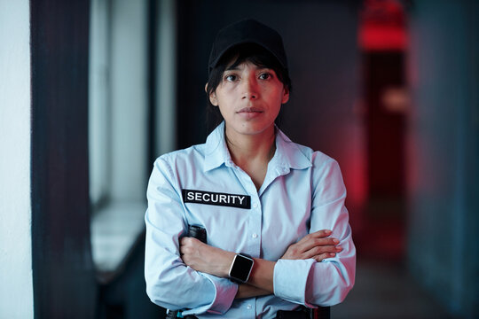 Young Hispanic female security guard in uniform looking at camera and crossing arms by chest while standing in dark corridor