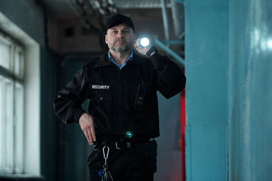 Mature male security guard in uniform and cap lighting forward with flashlight while moving along corridor of old building