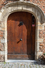 A historic wooden door with a stone arch is set in a brick building, with sunlight creating shadows...