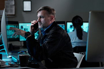 Side view of mature male security guard in black uniform sitting by workplace in surveillance room...