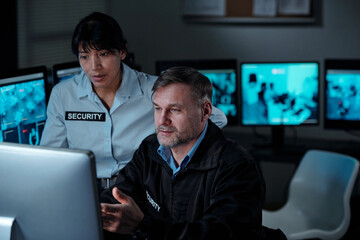Young female and mature male security guards in uniform looking at computer screen while one of...