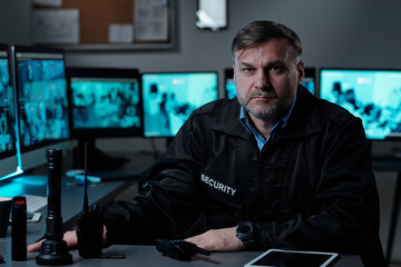 Serious and confident mature man in guard uniform looking at camera while sitting by workplace with computer monitors and transceivers