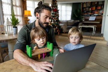 Fatherhood and remote business. Happy young single dad working at home while kids plays next to him