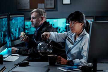 Young woman in uniform of security guard sitting by workplace in surveillance room and pouring tea in mug against mature man