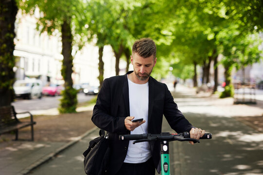 Businessman text messaging over mobile phone while standing with electric push scooter on footpath