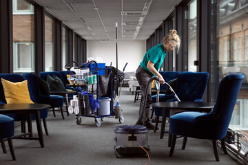 Full length of mature female janitor cleaning lobby with vacuum cleaner