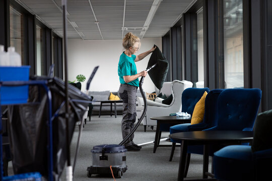 Mature female janitor using vacuum cleaner on cushion while cleaning lobby