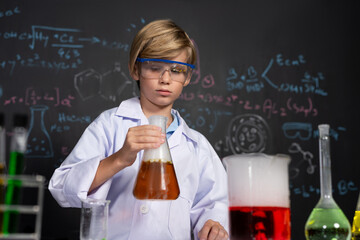 Blonde hair schoolboy in laboratory wear lab coat stand and learn science of chemistry technology in STEM class. The student hold beaker with orange liquid. On the table put many flask. Erudition.
