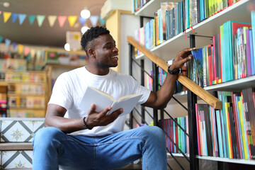 Young African student studying in a university library, holding a book, sitting by a bookshelf.