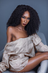 Portrait of beautiful Black woman with long curly black hair and beautiful makeup sitting by herself on lounge chair inside studio with grey background wearing brown silk dress shirt.