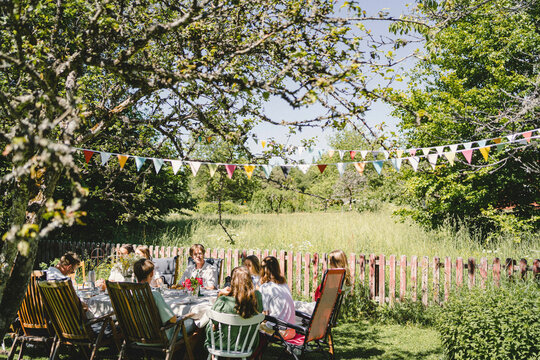 Young friends sitting at dining table under bunting decoration in back yard during summer solstice party