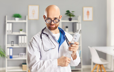 Portrait of funny young man doctor in white uniform with stethoscope holding big syringe in hands wearing glasses and looking at camera standing in medical clinic. Health care concept.