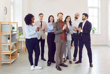 Obraz premium Group of company employees congratulating their happy male colleague on job well done applauding standing in office. Business people and group of staff clapping to young smiling man.