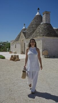 A young beautiful hispanic woman in white attire is walking happily near traditional trulli houses in alberobello, a historic old town in puglia, italy, on a sunny day.