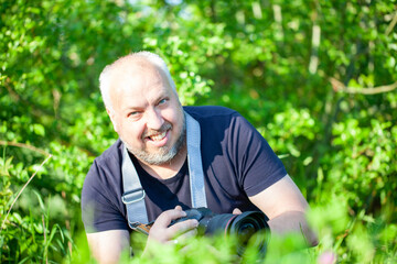A mature man with a camera is smiling while capturing outdoor photography in nature with a greenery...