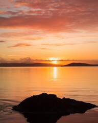 Naklejka premium A tranquil sunset over calm waters with a silhouette of a rock in the foreground and serene orange-pinkish skies reflecting on the water.