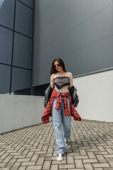 Cool fashionable stylish urban woman with sunglasses in fashion casual clothes with a leather jacket, bandana, jeans and shirt walks in the city near the building