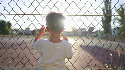 boy stands near the playground. concept of a happy childhood and loving family. a child holds on to the net of a sports ground, court and glare of lifestyle the sun in the background