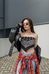 Stylish beautiful young urban woman model in fashionable clothes with leather jacket, jeans and bandana top puts on sunglasses and walks in the city