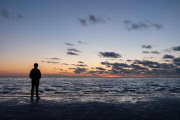 A person is on the beach, watching the sunset under a cloudy sky, with a tranquil sea spreading out before them