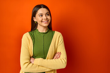 A brunette woman standing with arms crossed against an orange backdrop exuding confidence and poise.
