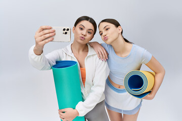 Two pretty teenage friends, sporting brunette hair and athletic wear, capture a selfie with a yoga mat in a studio setting.