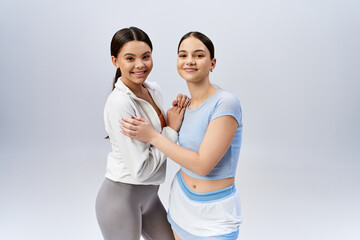 Two pretty, sportive teenage girls, one brunette, posing confidently together in a studio against a grey background.