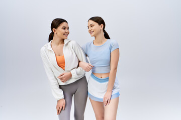 Two pretty, brunette young women in sportive attire stand next to each other, exuding confidence and friendship on a grey studio background.