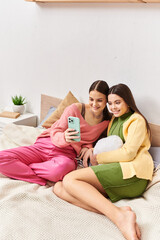 Two women, pretty teenage friends, sitting on a bed and engrossed in a cell phone screen.