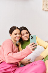 Two pretty teenage girls in casual attire sitting on a couch, capturing a fun moment by taking a selfie together.