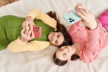 Two young women in casual clothing enjoy a moment of relaxation as they lay side by side on a cozy bed.