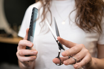 Hairdresser holding a comb and scissors in her hands standing in the salon. 