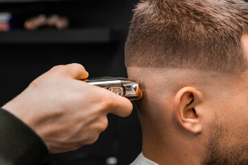 Barber uses shaver machine to cut young man hair in barbershop closeup. Master does modern haircut to male client in professional salon