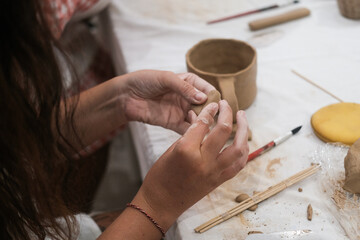 A close-up of the hands of a young woman as she shapes a heart out of clay to decorate a mug....