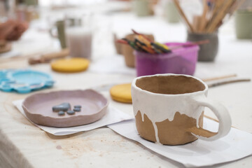 View of the table in a pottery workshop during a master class. On the table there is a mug and a plate made of clay by one of the beginning students. The products are decorated with colorful flowers.