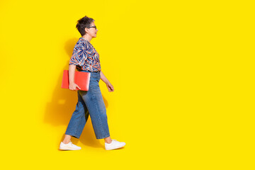 Full body photo of attractive retired woman hold netbook walk dressed stylish colorful clothes isolated on yellow color background