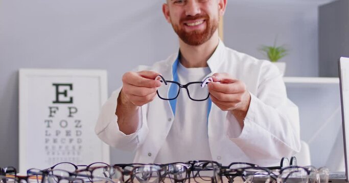 Smiling ophthalmologist or optician doctor sitting at desk in office with lot of eyeglasses offering glasses to patient looking at camera in clinic. Ophthalmology, eye health concept. 4k video.