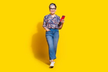 Full body photo of attractive retired woman walk hold device dressed stylish colorful clothes isolated on yellow color background