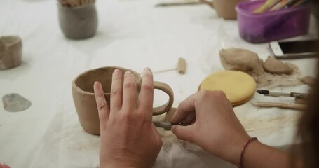 A close-up of the hands of a young woman who sculpts a mug from clay at a pottery master class. The process of creating a mug from clay at the beginner level in workshop. Creative hobby for adults.