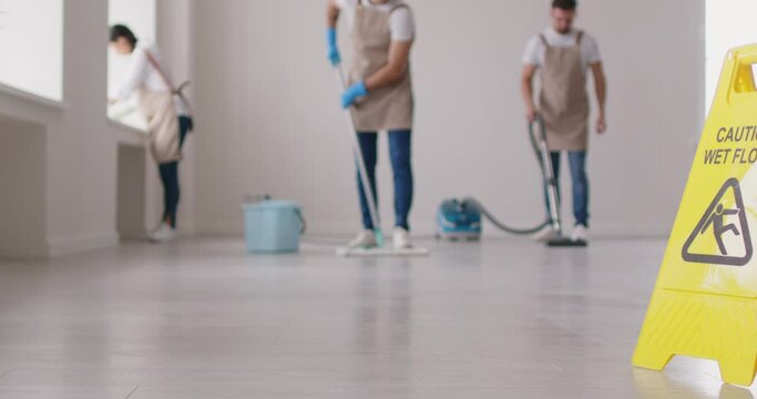 Camera slowly moves from left to right showing laminate surface and close up yellow plastic Caution Wet Floor sign while three cleaning service workers are mopping, vacuuming and wiping in backgroun