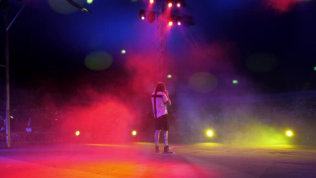 An outrageous circus presenter with a prosthetic leg and a microphone in his hands stands in the circus arena, under the light of colorful spotlights and smoke. A professional circus show.