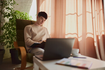 Young man, college student sitting on chair at home, making notes. Online studying course, blurred...