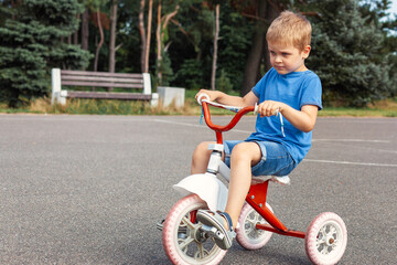 A little boy in blue clothes, very focused rides an red tricycle in city park