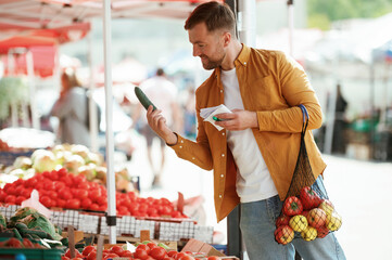 With a shopping list. Vegetables and fruits. Handsome man is on the street market or bazaar