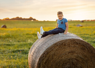 Portrait of a happy child against the background of rural fields in summer time, sitting on a roll...