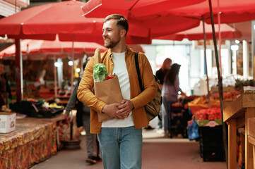 Smiling, holding shopping paper bag with products. Handsome man is on the street market or bazaar