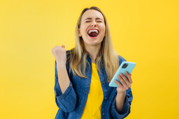 pretty girl hold smartphone amazed face open mouth scream yes win isolated on yellow color...