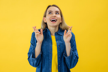 smiling young girl standing over yellow background, holding fingers crossed for good luck