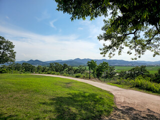 A path winds through a grassy field with a view of mountains in the distance. The sky is clear and blue, and the trees are lush and green. The scene is peaceful and serene, with a sense of calm