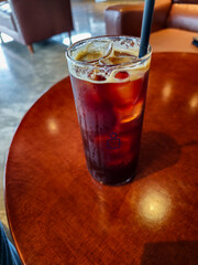 A glass of iced coffee sits on a wooden table. The glass is half full and the drink is a dark red color